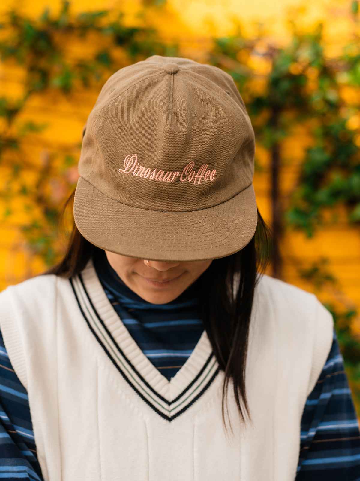 A photo of a person with their head tilted down while wearing an olive green hat with a pink embroidery that reads "dinosaur coffee"