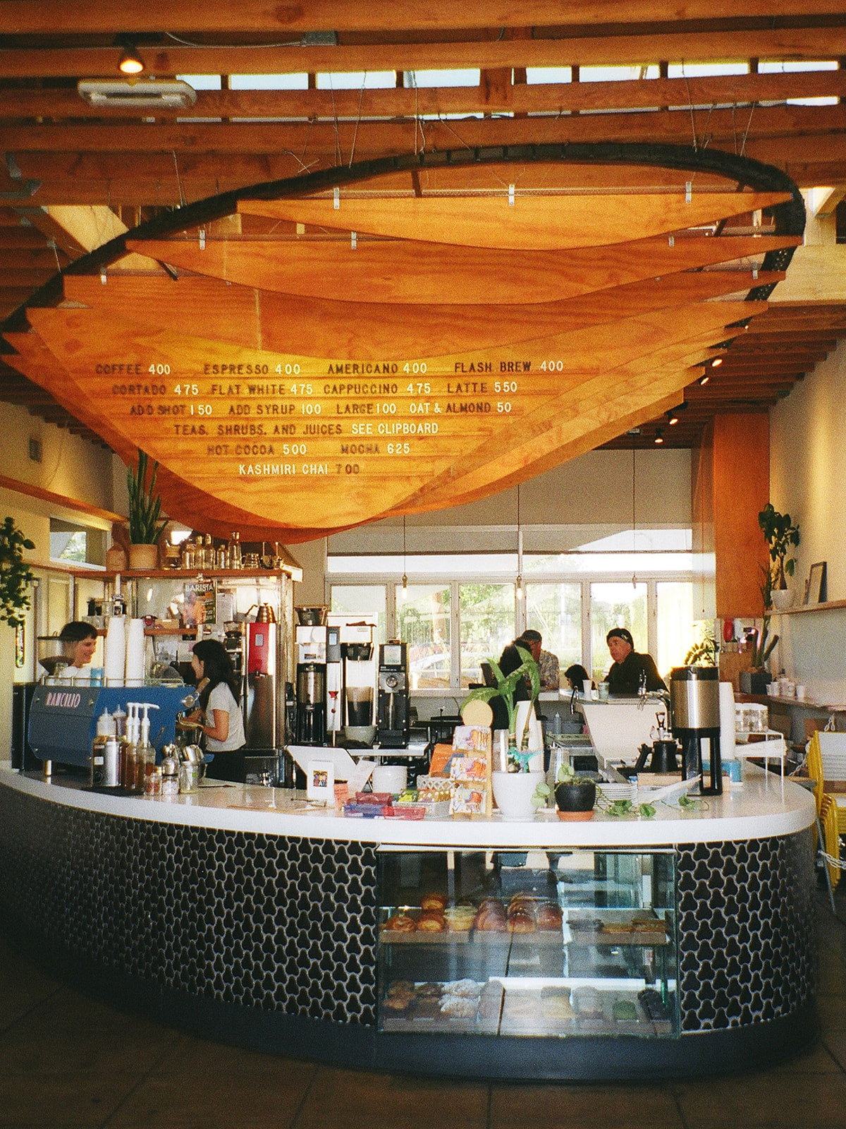 A photograph of a coffee shop interior. The ceiling is lined with cascading light wood panels, where the menu reads: coffee, espresso, americano, flash brew, cortado, flat white, cappuccino, latte, add shot, add syrup, kashmiri chai" The bar is lined with black tile in a simple brick pattern. Two female-identifying baristas laugh in the distant background"
