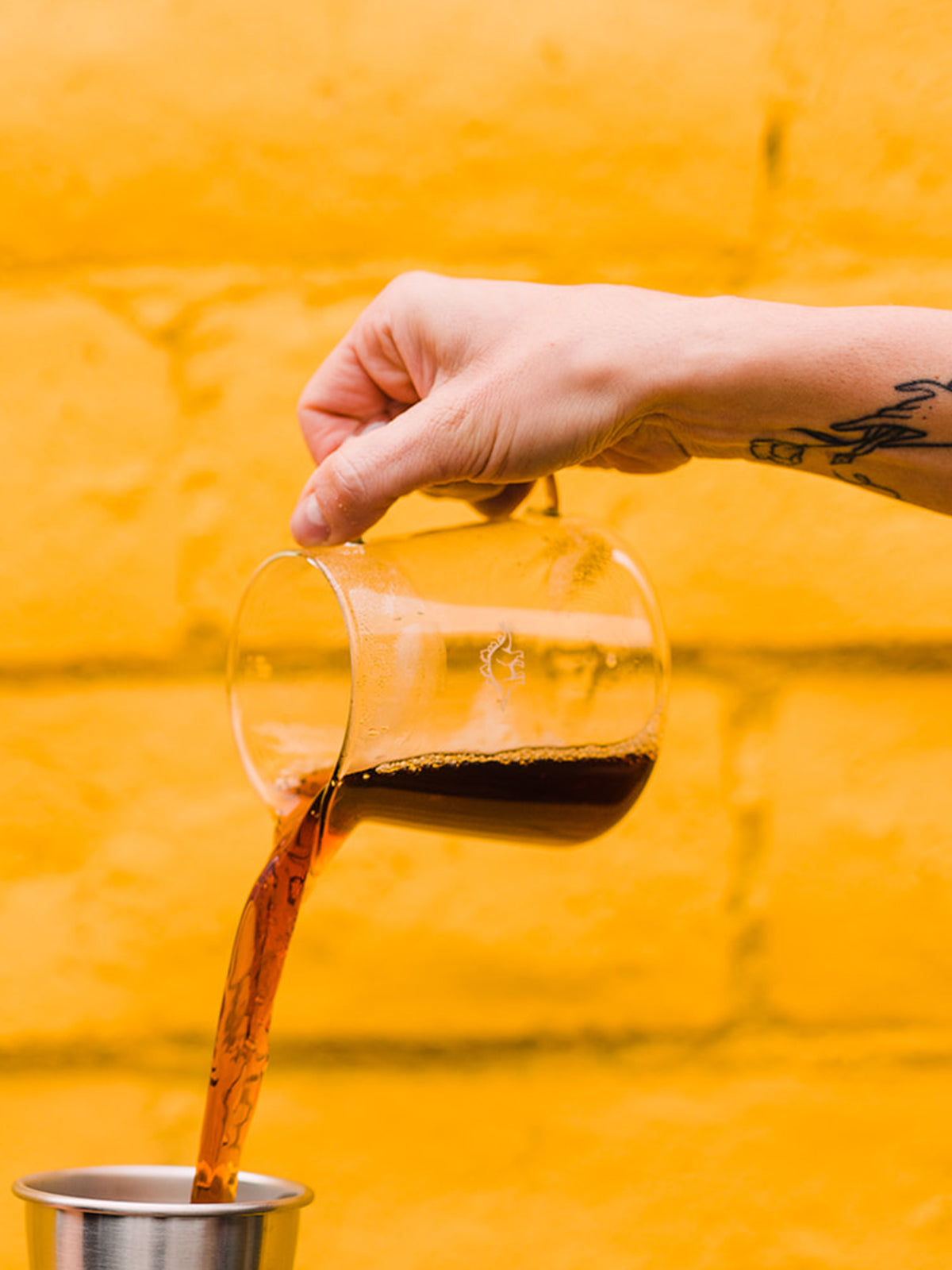 An out-of-frame hand pouring coffee from a clear pitcher into a tin cup 